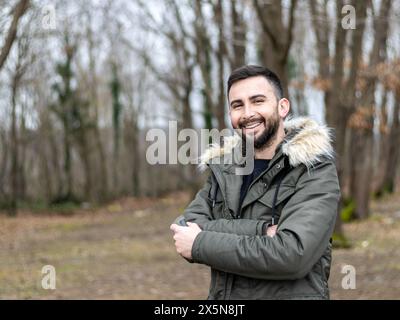 Junger und gutaussehender bärtiger Mann, der in die Kamera schaut und im Park, im Wald lächelt. Ein Mann, der bei kaltem Wetter einen Mantel trägt Stockfoto