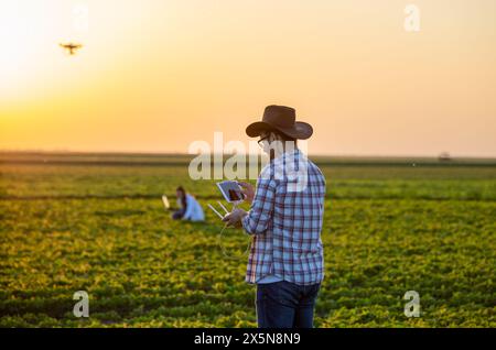 Landwirt, der bei Sonnenuntergang auf dem Feld steht und Drohne über Ackerland fährt, während Landwirtschaftsfrau im Hintergrund an einem Laptop arbeitet Stockfoto