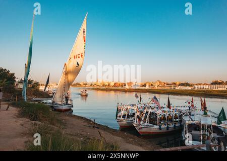 Eine Feluke und andere Touristenboote, die auf einer Bootstour bei Sonnenuntergang am Ufer des Nils in Luxor vor Anker gehen Stockfoto