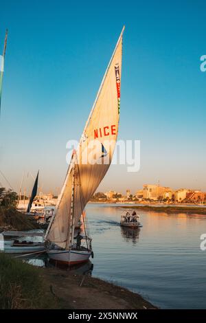Eine Feluke mit „SCHÖN“ gemalt auf dem Segel liegt bei Sonnenuntergang am Nil in Luxor, während ein weiteres Boot den Fluss unter einem klaren, goldenen Himmel überquert. Stockfoto