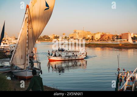 Während der goldenen Stunde segeln und docken die Boote entlang des Nil in Luxor an. Passagiere an Bord und Stadtgebäude leuchten im warmen Abendlicht über dem Riv Stockfoto