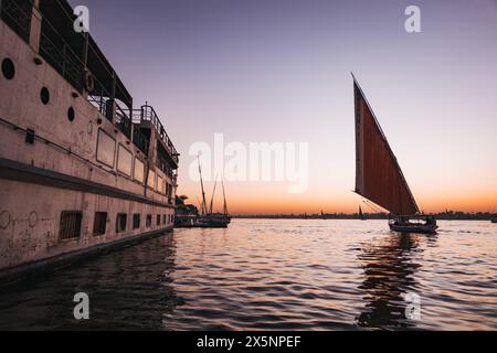 Eine Feluke segelt bei Sonnenuntergang an einem angedockten Kreuzfahrtschiff auf dem Nil vorbei, mit ruhigem Wasser und einem sanften orange-violetten Himmel über dem fernen Horizont. Stockfoto