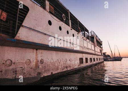 Seitenansicht eines alten Flussschiffes, das bei Sonnenuntergang am Nil angedockt ist, mit ruhigem Wasser und Segelbooten in der Ferne unter einem weichen lila und orangen Himmel. Stockfoto