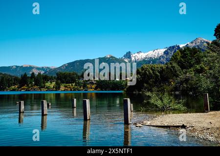 Felsiges Ufer mit grüner Vegetation in der Nähe eines blauen Sees, am Horizont eine Berglandschaft mit Häusern im Wald, Bariloche Nationalpark Stockfoto