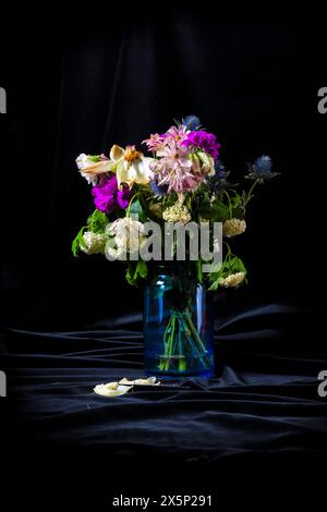 Eine Vase gefüllt mit verwelkten Blumen, auf schwarzem Hintergrund. Die Vase ist blau und enthält eine Vielzahl von Blumen, darunter lila und weiße. Die Stockfoto