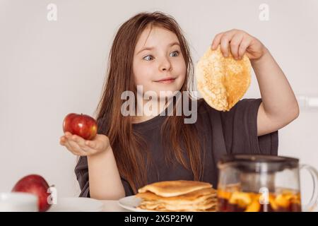 Ein kleines Teenager-Mädchen, das einen Apfel in der einen Hand in der anderen Pfannkuchen hält. Das Mädchen wählt zwischen einem Apfel und einem Pfannkuchen zum Frühstück, sieht aus, als ob es ihn leckt Stockfoto