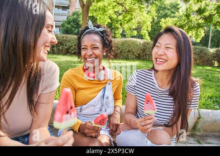 Drei junge Multi-ethnische nette Frauen, die Spaß beim Eishalten haben, sitzen draußen im Sommerpark. Stockfoto