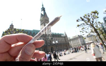 Ein Mann hält einen Joint in der Hand. Im Hintergrund das Hamburger Rathaus. Der Hamburger Senat hat einen Katalog von Bußgeldern für Gesetzesverstöße erlassen, der hohe Geldstrafen und sogar Haftstrafen für Personen vorsieht, die mehr als die erlaubten 25 Gramm Haschisch oder Marihuana auf der Straße dabeihat oder 50 Gramm in der Wohnung besitzt. Altstadt Hamburg *** Ein Mann hält einen Joint in der Hand Hamburger Rathaus im Hintergrund hat der Hamburger Senat einen Katalog von Bußgeldern für Gesetzesverstöße herausgegeben, der hohe Bußgelder und sogar Freiheitsstrafen für Personen vorsieht, die mehr tragen Stockfoto