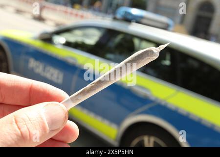 Ein Mann hält einen Joint in der Hand. Im Hintergrund ein Einsatzwagen der Hamburger Polizei. Der Hamburger Senat hat einen Katalog von Bußgeldern für Gesetzesverstöße erlassen, der hohe Geldstrafen und sogar Haftstrafen für Personen vorsieht, die mehr als die erlaubten 25 Gramm Haschisch oder Marihuana auf der Straße dabeihat oder 50 Gramm in der Wohnung besitzt. Altstadt Hamburg *** Ein Mann hält einen Joint in der Hand Ein Hamburger Polizeiauto im Hintergrund der Hamburger Senat hat einen Katalog von Bußgeldern für Gesetzesverstöße herausgegeben, der hohe Bußgelder und sogar Freiheitsstrafen für Menschen vorsieht Stockfoto