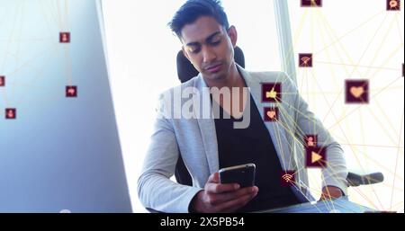 Bild von vernetzten Ikonen über birassischen Männern, die auf einem Stuhl sitzen und im Büro ein Mobiltelefon benutzen Stockfoto