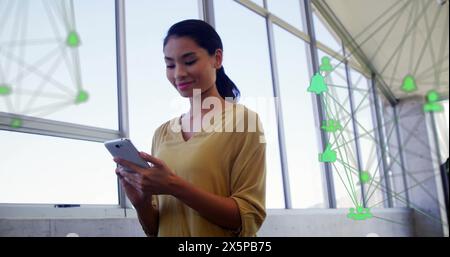 Bild von verbundenen Ikonen über lächelnder asiatischer Frau, die im Büro auf dem Smartphone scrollt Stockfoto