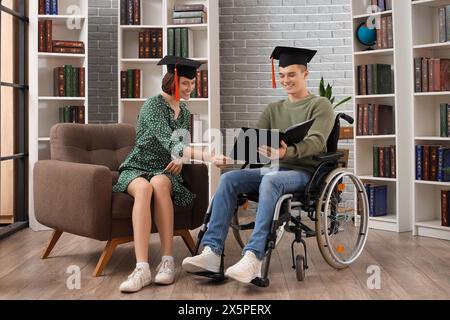 Männlicher Rollstuhlabsolvent mit Buch und sein Klassenkamerad in der Bibliothek Stockfoto