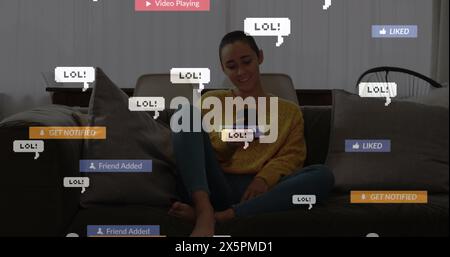Bild von Social-Media-Ikonen über birassische Frau, die lächelt, während sie ihr Smartphone zu Hause verwendet Stockfoto