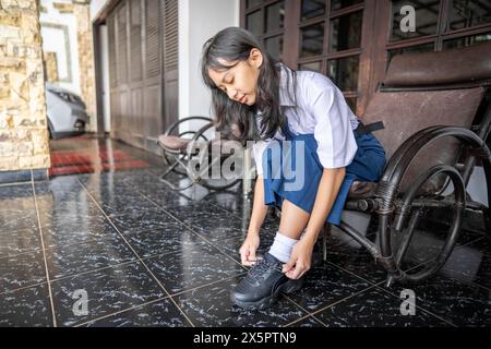 Junge Indonesin mit Schuluniform, die morgens vor dem Haus zur Schule geht und Schnürsenkel bindet. Back-to-School-Konzept. Stockfoto