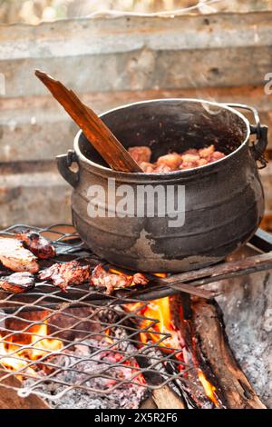 afrikanische Küche, Fleisch auf dem Grill, großer schwarzer Kessel, der mit Eintopf im Hintergrund kocht Stockfoto