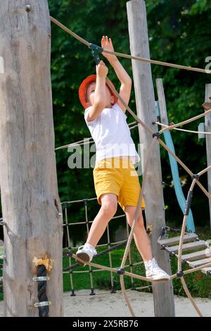 Ein Kind klettert an einem heißen Sommertag in einem Park auf einem Spielplatz auf ein alpines Netz. Kinderspielplatz in einem öffentlichen Park, Unterhaltung und Erholung f Stockfoto