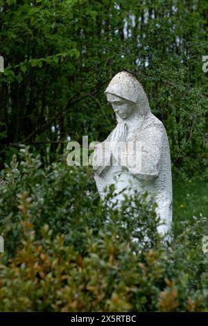 Eine beschädigte, alte Statue der Mutter Gottes, verlassen in der Friedhofskippe, verwischte Bäume mit grünen Blättern im Hintergrund. Stockfoto