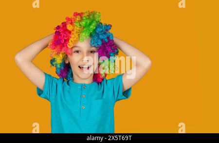 Lustiger Junge in mehrfarbiger Perücke. Lächelnder kleiner Junge in Clown-Perücke. Glücklicher Clown Junge mit großer bunter Perücke - isoliert auf gelbem Hintergrund. Niedliches Kind Stockfoto