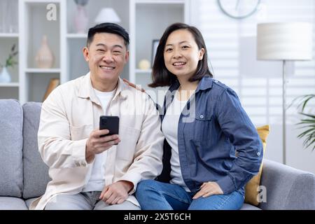 Ein fröhliches Paar mittleren Alters genießt einen entspannten Moment auf dem Sofa im Wohnzimmer. Der Mann hält ein Smartphone, beide lächeln warm. Sie sind lässig gekleidet Stockfoto