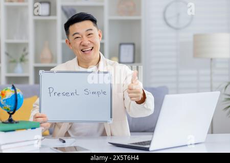 Der asiatische Lehrer gibt gerne einen Daumen hoch, während er online die gegenwärtige einfache Form unterrichtet. Helles Home Office mit Laptop, Büchern und einem Globus, das eine ansprechende und lehrreiche Umgebung schafft. Stockfoto