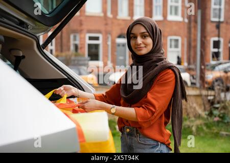 Frau im Hijab, die Einkaufstaschen aus dem Kofferraum auslädt, Porträt Stockfoto