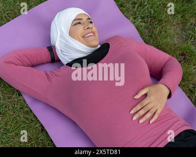 UK, Sutton, lächelnde Frau im Kopftuch, die auf Yogamatte auf Gras liegt Stockfoto