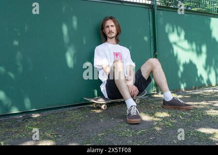 Porträt eines jungen Mannes, der im Park auf dem Skateboard sitzt Stockfoto