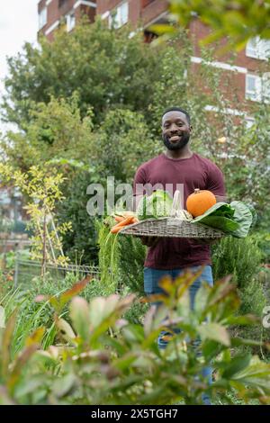 Porträt eines lächelnden Mannes, der Korb mit frischem Gemüse im städtischen Garten hält Stockfoto