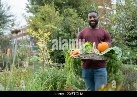 Porträt eines lächelnden Mannes, der Korb mit frischem Gemüse im städtischen Garten hält Stockfoto