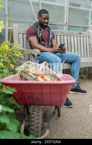 Mann mit Smartphone, der auf einer Bank im Kleingarten sitzt Stockfoto