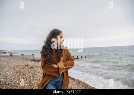 Nachdenkliche Frau, die auf das Meer blickte, während sie am Strand stand, an bewölktem Tag Stockfoto