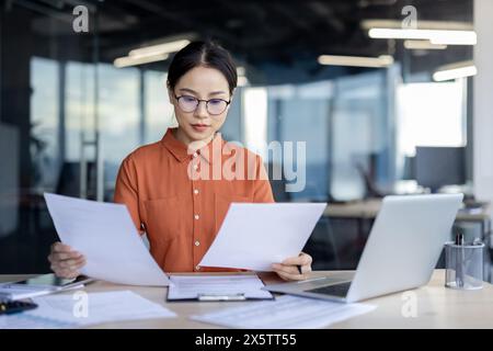 Eine ernstzunehmende junge professionelle Frau in einem stylischen orangefarbenen Hemd bewertet Papierkram aufmerksam, umgeben von Laptops in einer gut beleuchteten Büroumgebung. Ihr Fokus und ihre Entschlossenheit sind offensichtlich. Stockfoto