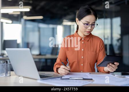 Ernsthafte junge asiatische Geschäftsfrau konzentrierte sich auf die Prüfung von Finanzdokumenten und hielt einen Taschenrechner in moderner Büroumgebung. Stockfoto