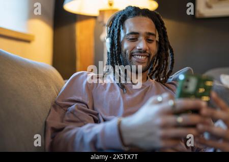 Mann mit Dreads, der das Telefon benutzt und Musik hört, während er sich auf dem Sofa entspannt Stockfoto