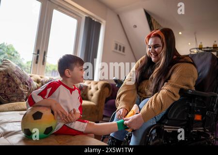 Frau im Rollstuhl bindet Söhne in Fußballschuhen im Wohnzimmer Stockfoto