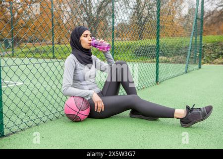 Frau im Hijab mit Basketballball, die auf dem Basketballfeld sitzt Stockfoto