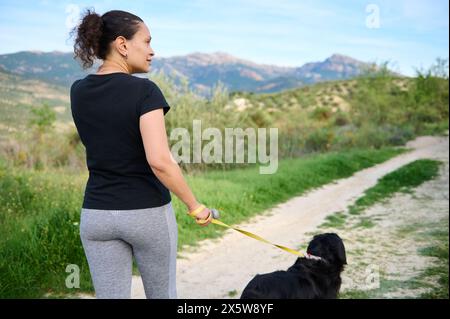 Blick von der Rückseite einer jungen, sportlichen Frau in grauen Leggings und schwarzem T-Shirt, die ihren Hund auf einen Spaziergang mitnimmt, während sie in der Natur der Berge joggen Stockfoto