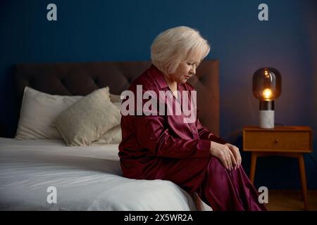 Ältere Frau im Pyjama, die auf dem Bett sitzt und Einsamkeit spürt Stockfoto