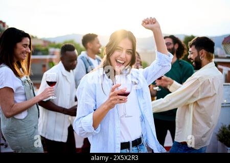 Junge weiße schöne Frau posiert mit Wein und Freunden, die auf dem Sommerdach tanzen. Stockfoto