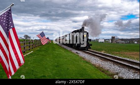 Ein Zug fährt die Gleise entlang, neben einem Feld mit amerikanischer Flagge. Der Zug ist schwarz-weiß und zieht einen Pkw. Die Fahnen winken im Wind Stockfoto