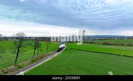 Dramatische Luftaufnahme einer alten Dampfeisenbahn, die auf einer kurvigen Strecke durch üppige grüne Felder fährt, vor dem Hintergrund eines dramatischen bewölkten Himmels und eines fernen Dorfes. Stockfoto