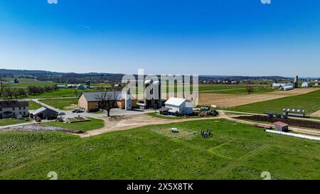 Ein Bild über der Decke eines belebten Bauernkomplexes mit mehreren Scheunen und Silos umgeben von grünen Weiden, in Betrieb befindlichen landwirtschaftlichen Geräten und einer Gruppe von Menschen, die sich unter klarem Himmel versammelt haben. Stockfoto