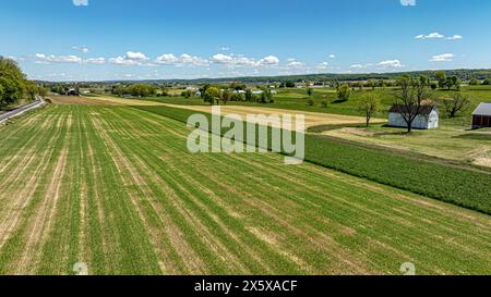 Ein Luftbild fängt die ausgedehnten und vielfältigen landwirtschaftlichen Felder ein und zeigt Muster verschiedener Kulturen, ein kleines Lagergebäude und eine ferne Eisenbahn unter einem Himmel voller flauschiger Wolken. Stockfoto