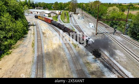 Ein leistungsstarker Oldtimer-Dampfzug fährt durch einen Industriebahnhof und emittiert Dampf, wenn er durch Lagerhäuser und neben anderen Eisenbahnwaggons fährt Stockfoto