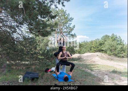 Drei Freunde üben Acro-Yoga-Übungen beim Wandern im Wald. Ein Mann, der auf dem Rücken liegt, hält zwei Mädchen an den Füßen. Stockfoto