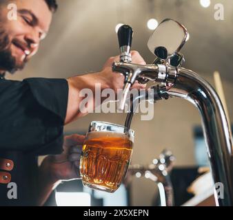 Ein lächelnder, stilvoller Bärenbarmann in schwarzer Uniform mit einer Schürze, der frisches Lagerbier in einen Glasbecher an der Bar zapft. Erfolgreiche Leute, Bierkonzern Stockfoto