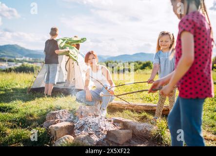 Drei Schwestern machen Picknick, rösten Marshmallows und Süßigkeiten auf Stöcken über Lagerfeuer, während zwei Brüder das grüne Zelt aufbauten. Glückliche Familie, Outd Stockfoto