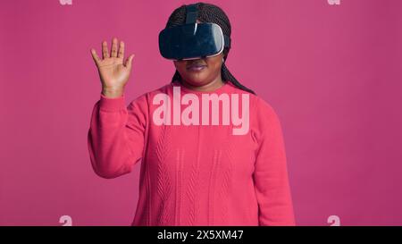 Die innovative junge schwarze Frau genießt futuristische Simulation mit interaktivem Gerät, während sie im Studio eine 3D-Virtual-Reality-Brille trägt. Trendige afrikanische Fashionista mit modernem VR-Headset. Stockfoto