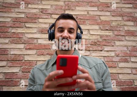 Nahaufnahme attraktiver junger Mann mit rotem Handy, das gegen eine Ziegelwand gelehnt ist. Süchtig machende Technologie. Stockfoto