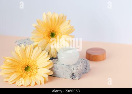 Kosmetische Creme auf Steinpodium mit gelben Gerbera-Blüten auf beigefarbenem Tisch. Nahaufnahme. Stockfoto